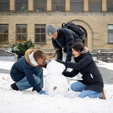 Three Cornellians are joyfully building a snowman on a snowy lawn in front of a charming stone building, capturing the essence of winter at Cornell University.