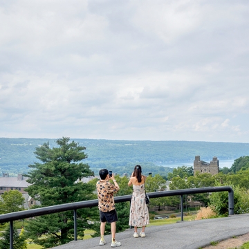 Two people stand on a pathway overlooking a scenic landscape with lush greenery and distant buildings, one person appears to be taking a photo of the other who is posing. A cloudy sky and a large expanse of water are visible in the background.