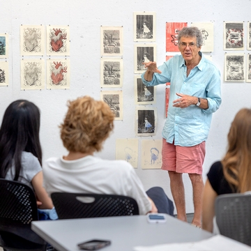 A professor standing and gesturing to a wall filled with art, during a printmaking class. A group of seated students, seen from the back, listens attentively.