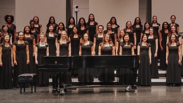 A large group of chorus members stand on stage behind a grand piano.
