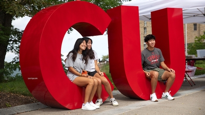 Three students sit on large letters of Cornell's intials 